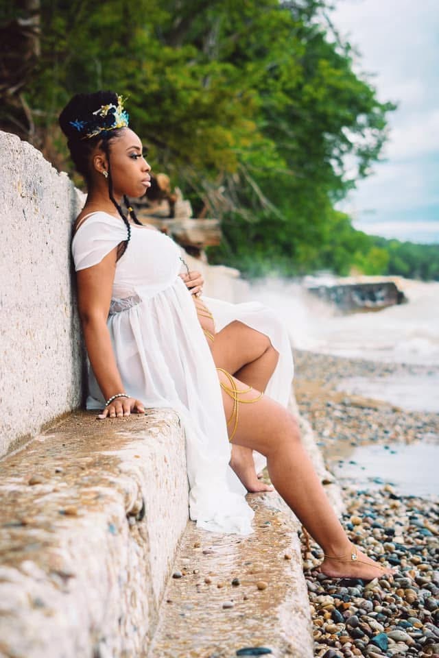 Omi seated against a stone wall by the shoreline during pregnancy, in a white gown and crown