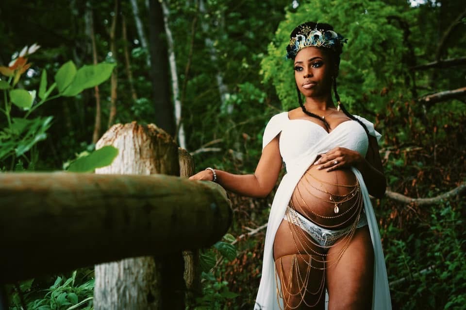 Expectant mother standing in a wooded clearing in a white gown, floral crown and gold belly adornment